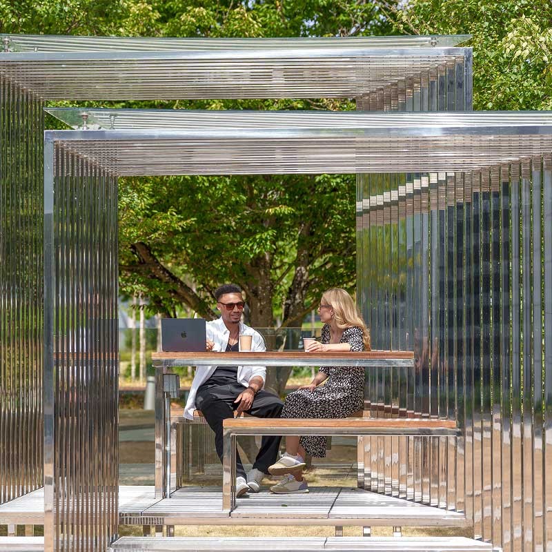 Two people sit and talk at a metal outdoor table structure with a laptop and drinks, surrounded by greenery and sunlight.