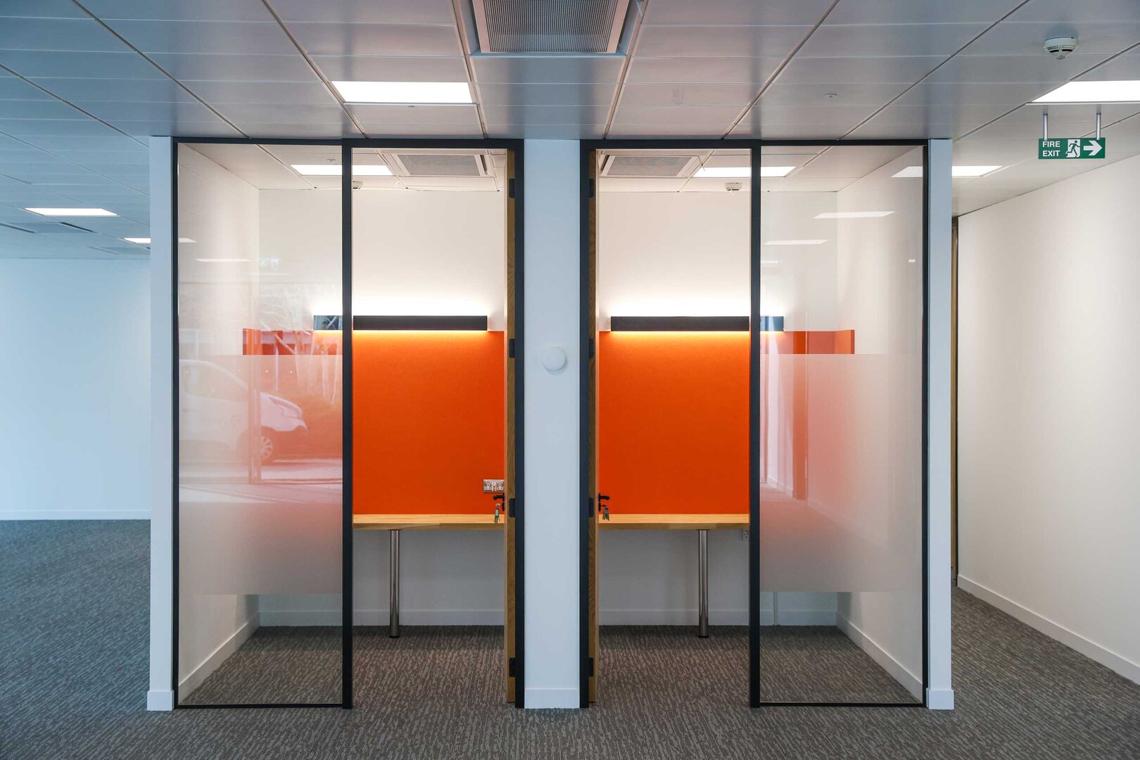Two glass-walled office pods with orange back panels and wooden desks, separated by a central wall, in a modern workplace.