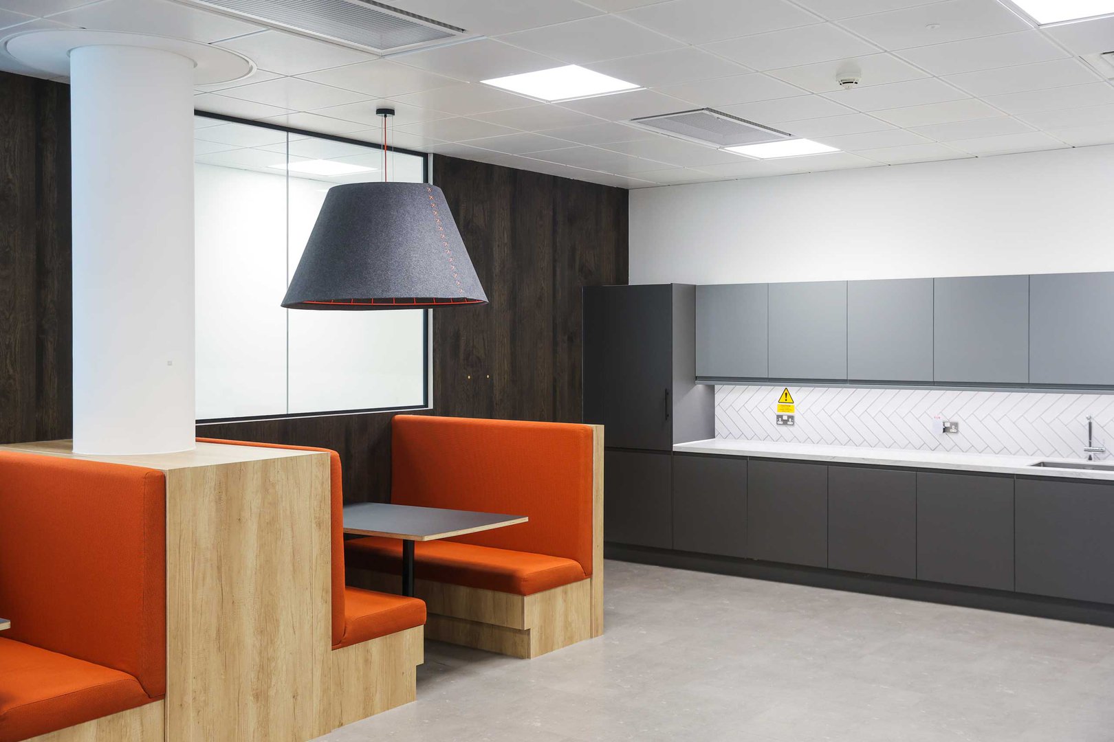 Modern office kitchen with grey cupboards, white splashback, sink, and orange booth seating around a table under a large pendant light.