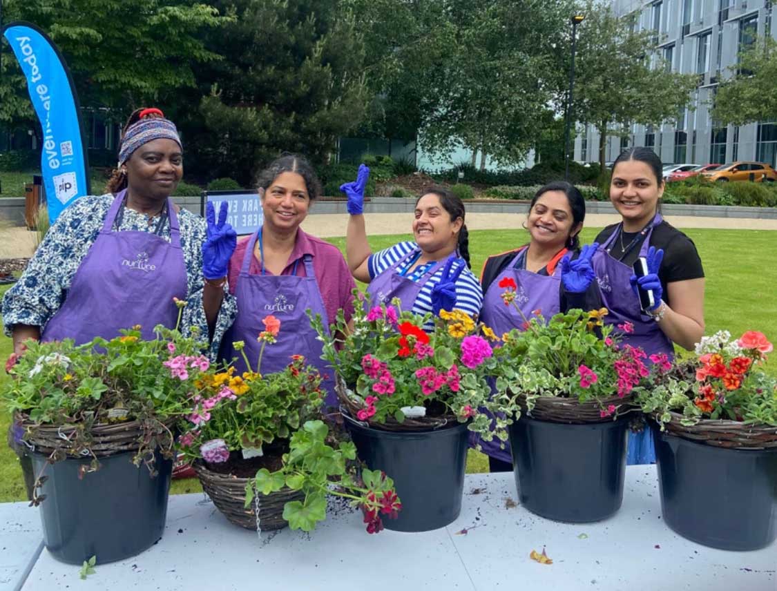 Five women in purple aprons and blue gloves stand outside behind a table with baskets and pots of colourful flowers, smiling at the camera.