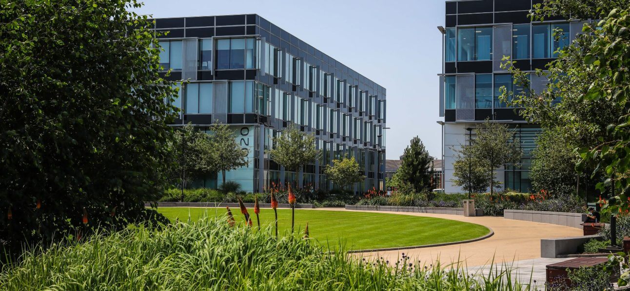 Two modern office buildings with large glass windows face a landscaped courtyard with a curved path, green lawn, and trees under a clear sky.