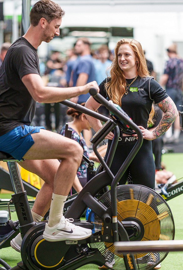 A man uses an air bike while a woman, likely a gym trainer, stands beside him, observing and smiling. Other people are visible in the background in a gym setting.