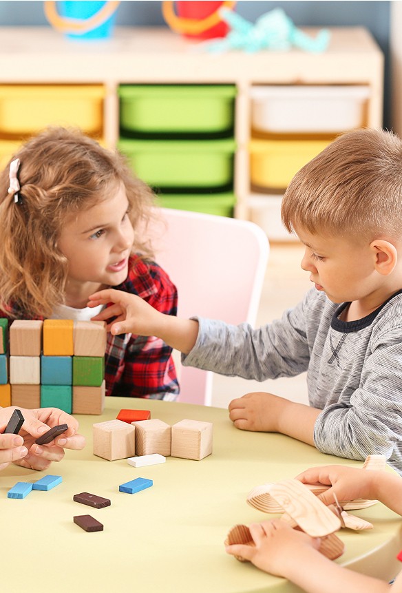 Two young children sit at a table playing with wooden blocks, whilst other children's hands are also visible interacting with toys in the foreground.