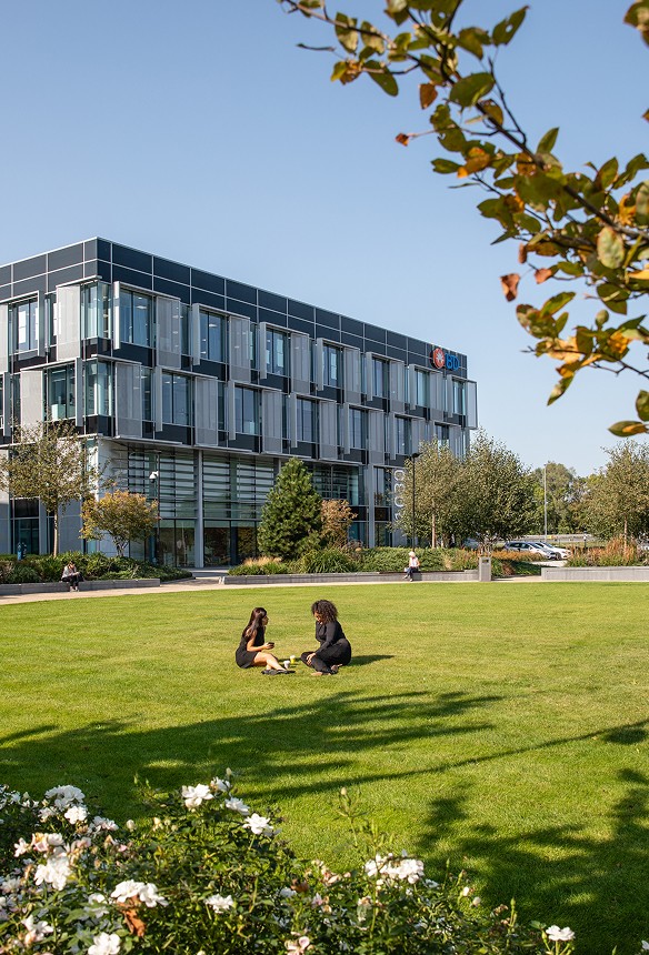 Two people sit on a grassy lawn in front of a modern glass building with the SGS logo on a sunny day, surrounded by trees and landscaping.