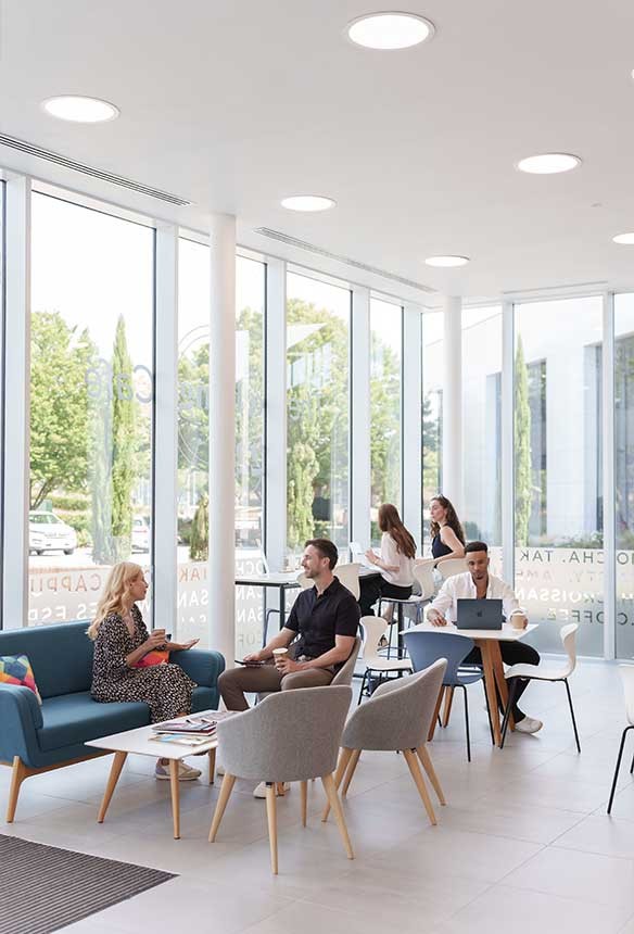 Modern office lounge with large windows, people sitting and talking, some working on laptops, natural light filling the space, and greenery visible outside.