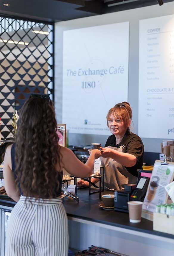 A barista hands a card to a customer across the counter at The Exchange Café, with a menu visible in the background.