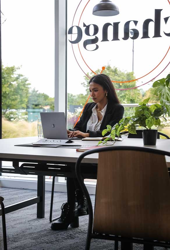 A woman in business attire works on a laptop at a desk in a modern office with plants and large windows.