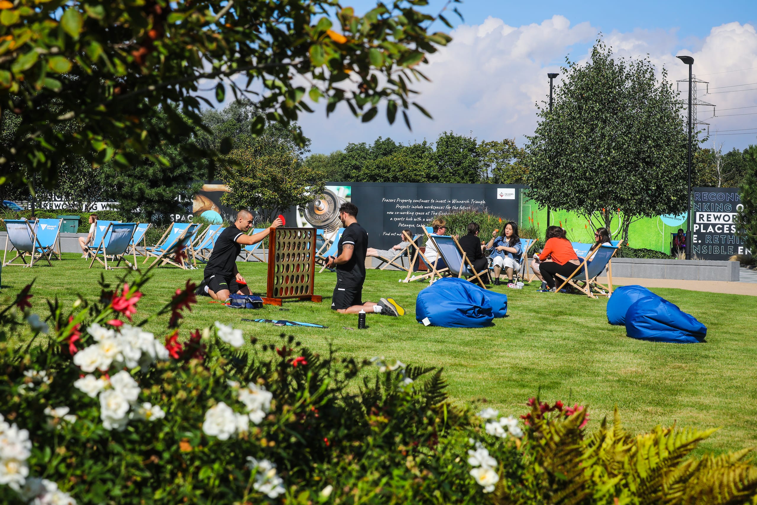 People play a large Connect Four game and relax on deckchairs and beanbags on a grassy outdoor area with flowers and trees.