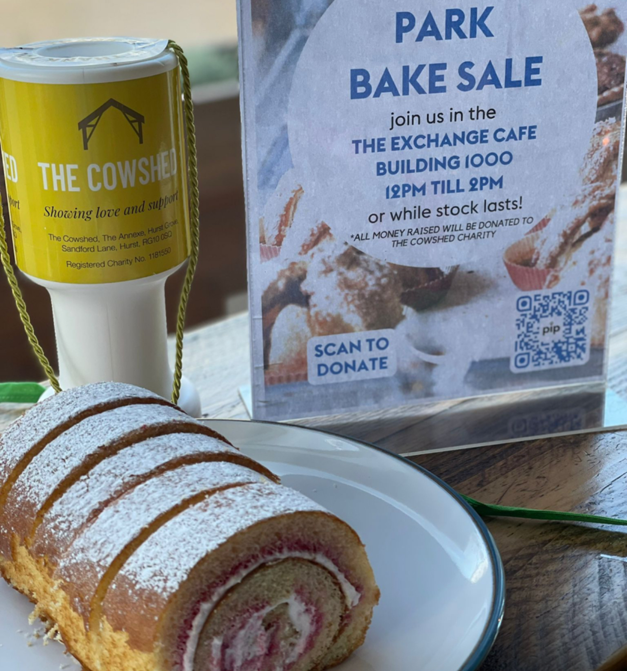 A Swiss roll on a plate sits next to a donation tin and a sign advertising a park bake sale benefitting The Cowshed charity.