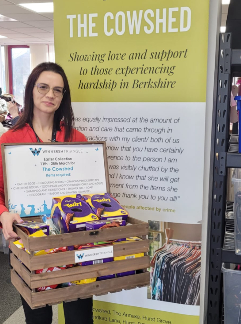 A woman stands holding a crate of Easter eggs in front of a banner for The Cowshed, an organisation supporting those experiencing hardship in Berkshire.