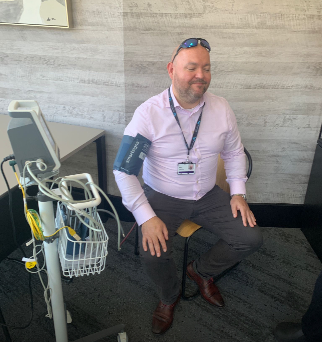 A man sits on a chair with a blood pressure cuff on his arm, connected to a monitor on a nearby stand. He is smiling with his eyes closed.