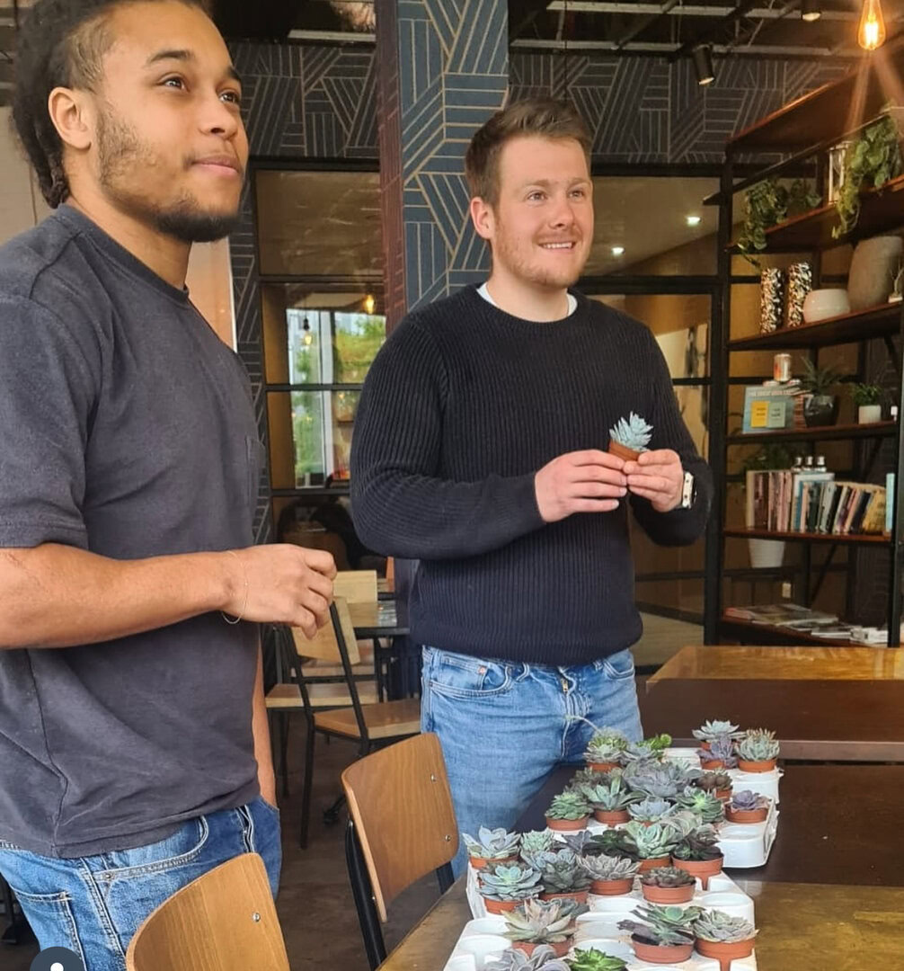 Two men stand indoors, holding small potted succulents, with more succulents arranged on a table in front of them. The setting appears to be a workshop or café.