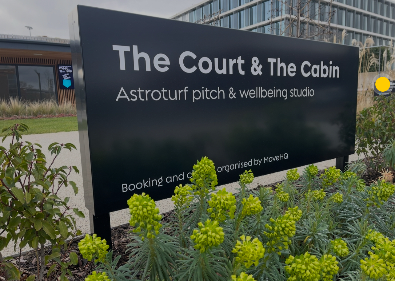 A black sign reads The Court & The Cabin, Astroturf pitch & wellbeing studio. Booking and classes organised by MoveHQ, with green plants in the foreground.