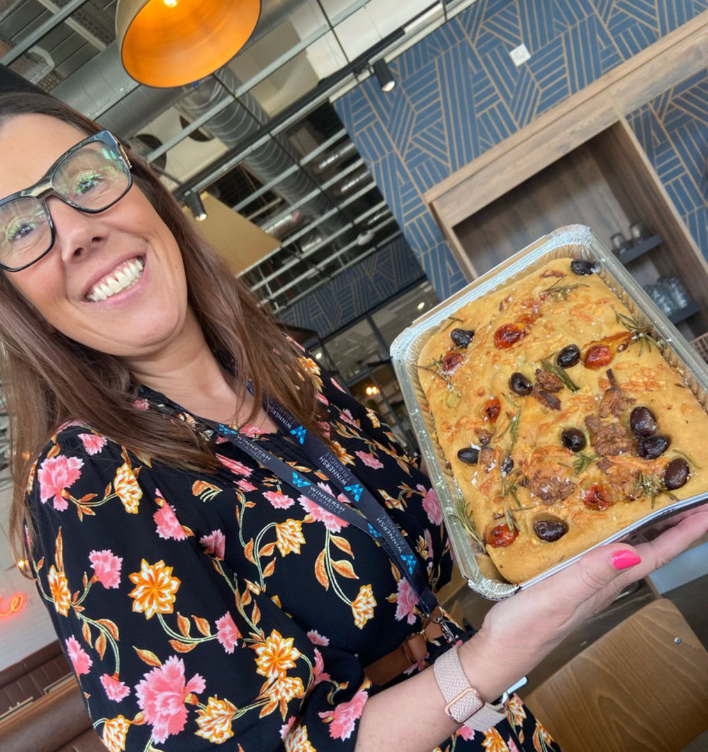 A woman wearing glasses and a floral dress holds a tray of focaccia bread topped with tomatoes, olives, and herbs inside a modern café.