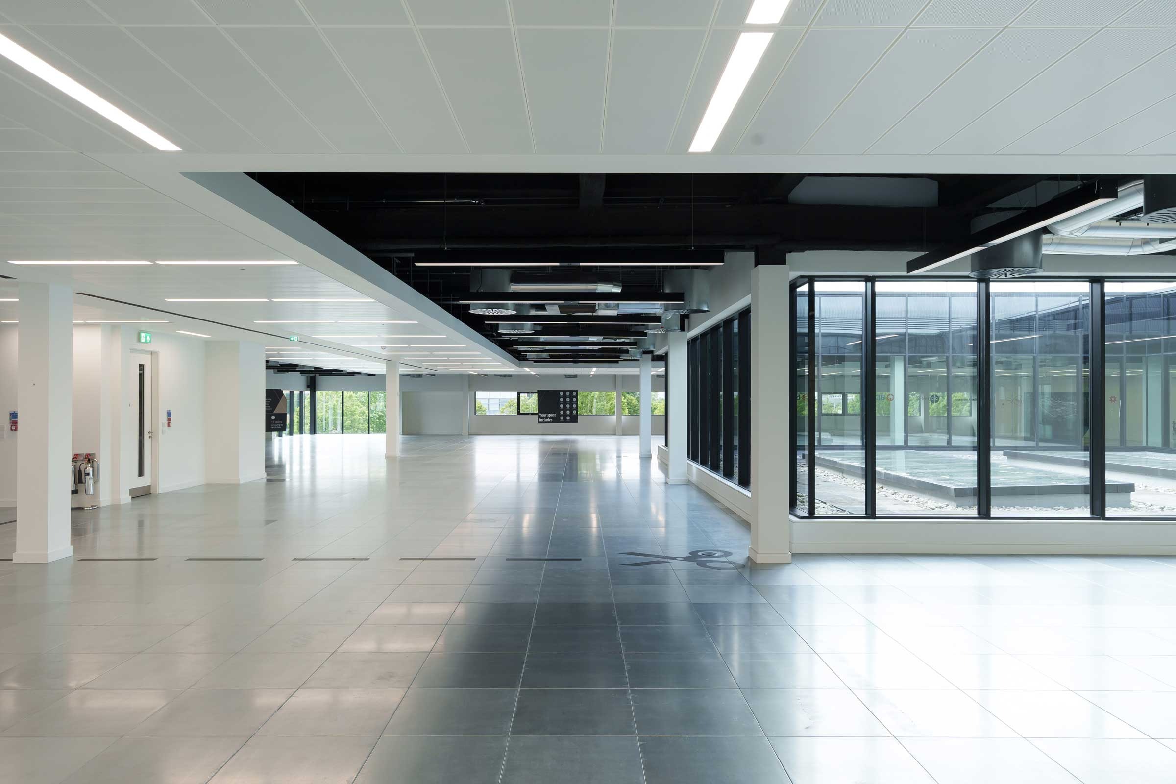 Spacious, modern, empty office interior with tiled floors, white walls, large windows, and overhead lighting; glass wall reveals courtyard area outside.