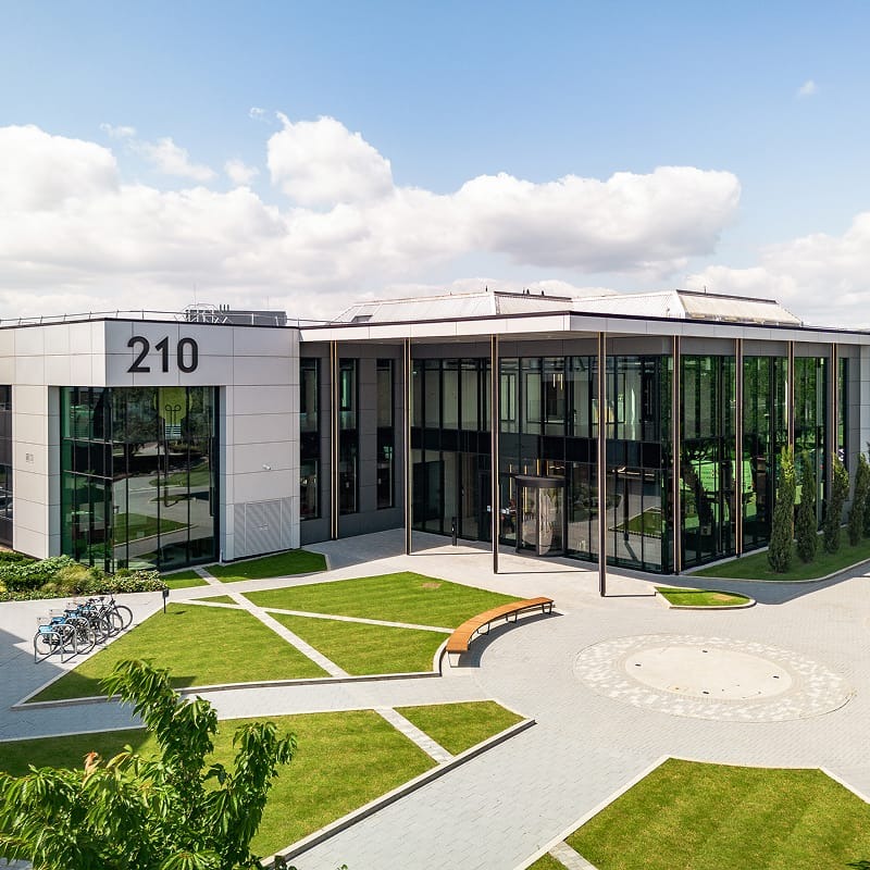 Modern office building with large windows and the number 210 on the exterior, surrounded by landscaped lawns, paths, benches, and parked bicycles under a partly cloudy sky.