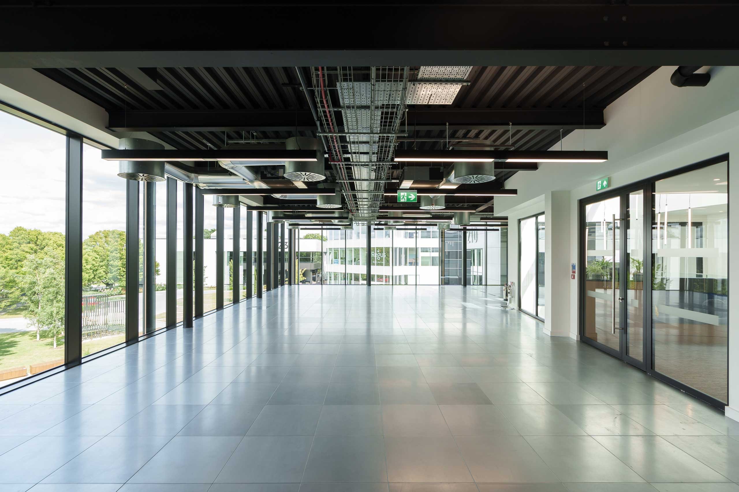 A modern, empty corridor with glass walls, tiled floor, exposed ceiling pipes, and exit signs, overlooking an outdoor area with trees and a white building.