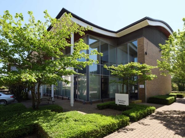 Modern office building with large glass windows, brick exterior, and landscaped greenery; a sign labelled Entrance is visible near the door.