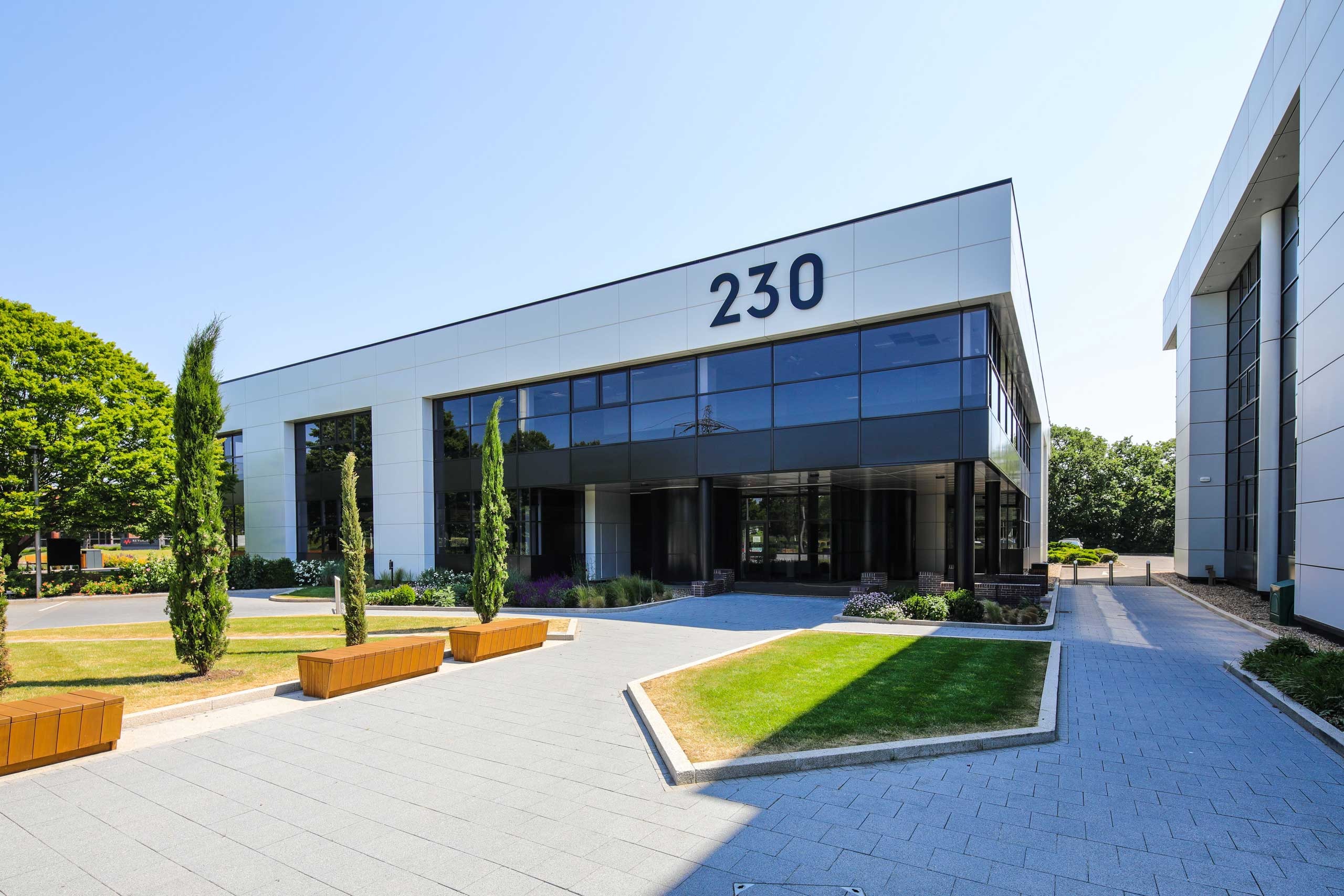 Modern office building labelled 230 with large windows, neatly landscaped grounds, and a paved walkway in the foreground on a clear day.