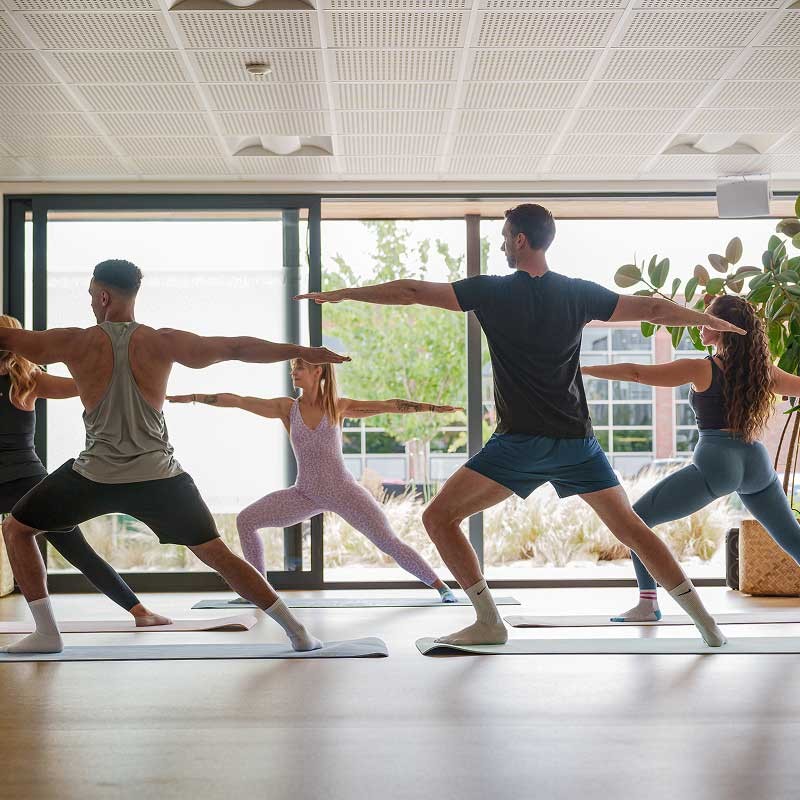 A group of people practise yoga indoors, standing on mats in a warrior pose, with large windows and natural light in the room.
