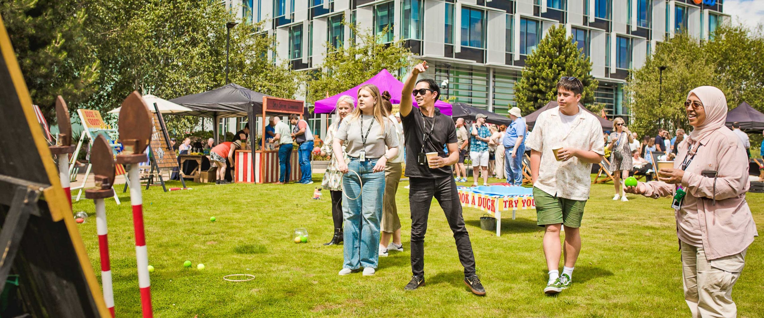 A group of people play outdoor games on a grassy lawn at an event, with marquees, stalls, and a modern building in the background.