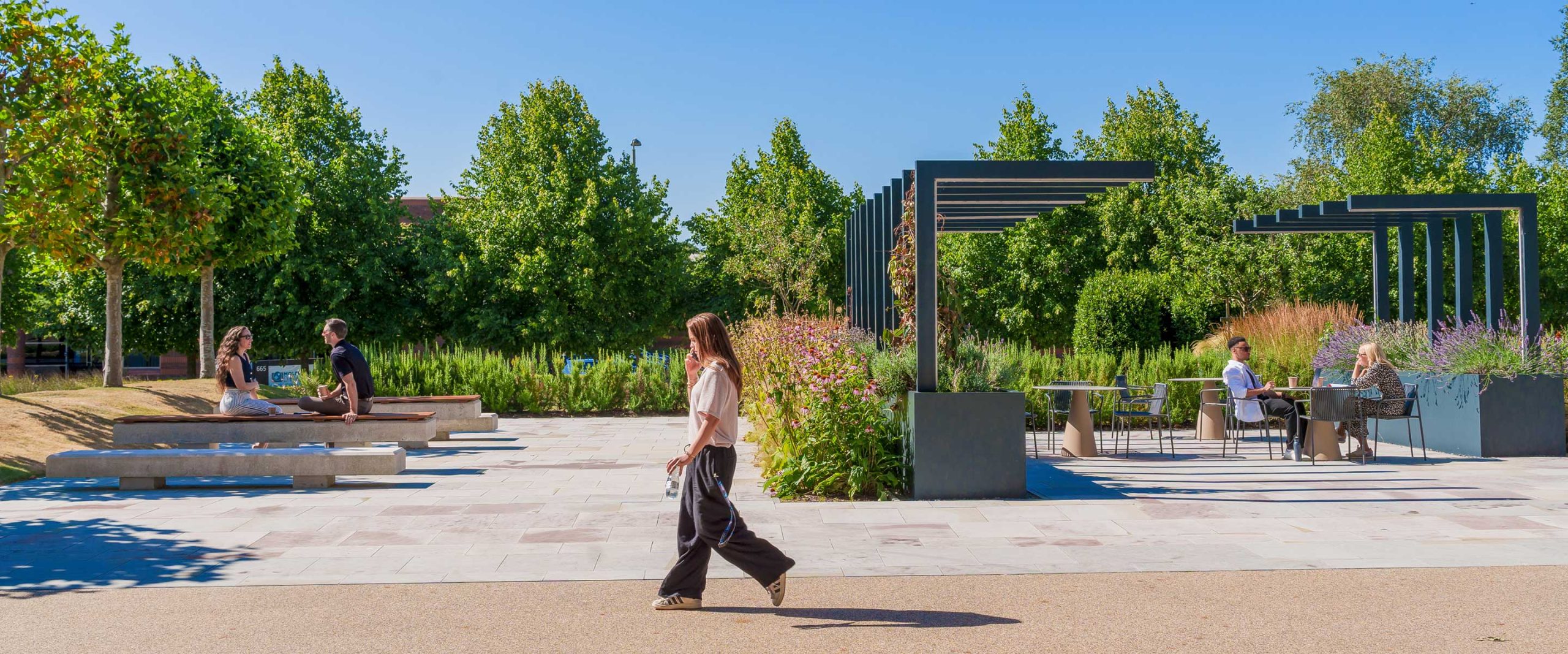 People sit and talk at tables and benches under pergolas in a landscaped outdoor park, whilst a woman walks by on a sunny day.