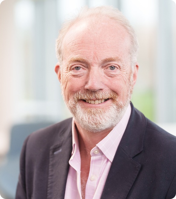 An older man with a white beard and light hair, wearing a dark blazer and light pink shirt, smiles at the camera in a brightly lit indoor setting.