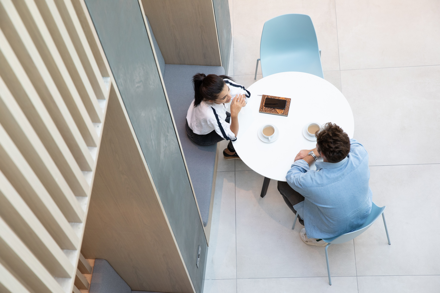 Two people sit at a round table with two cups and a tablet, viewed from above in a modern, minimalist setting.