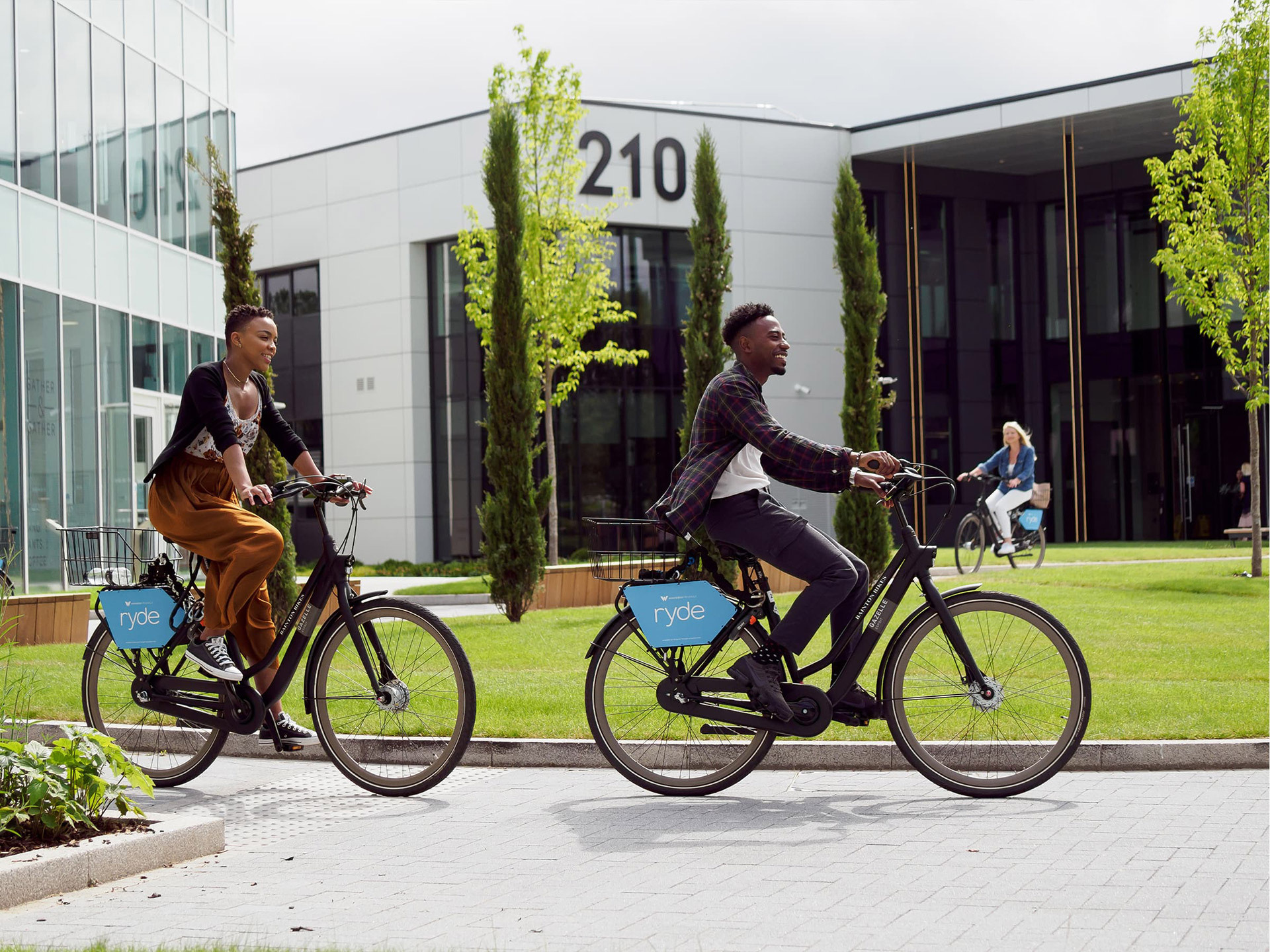 Two people ride hired Ryde bicycles on a pathway near a modern office building with a 210 sign, while another cyclist is visible in the background.