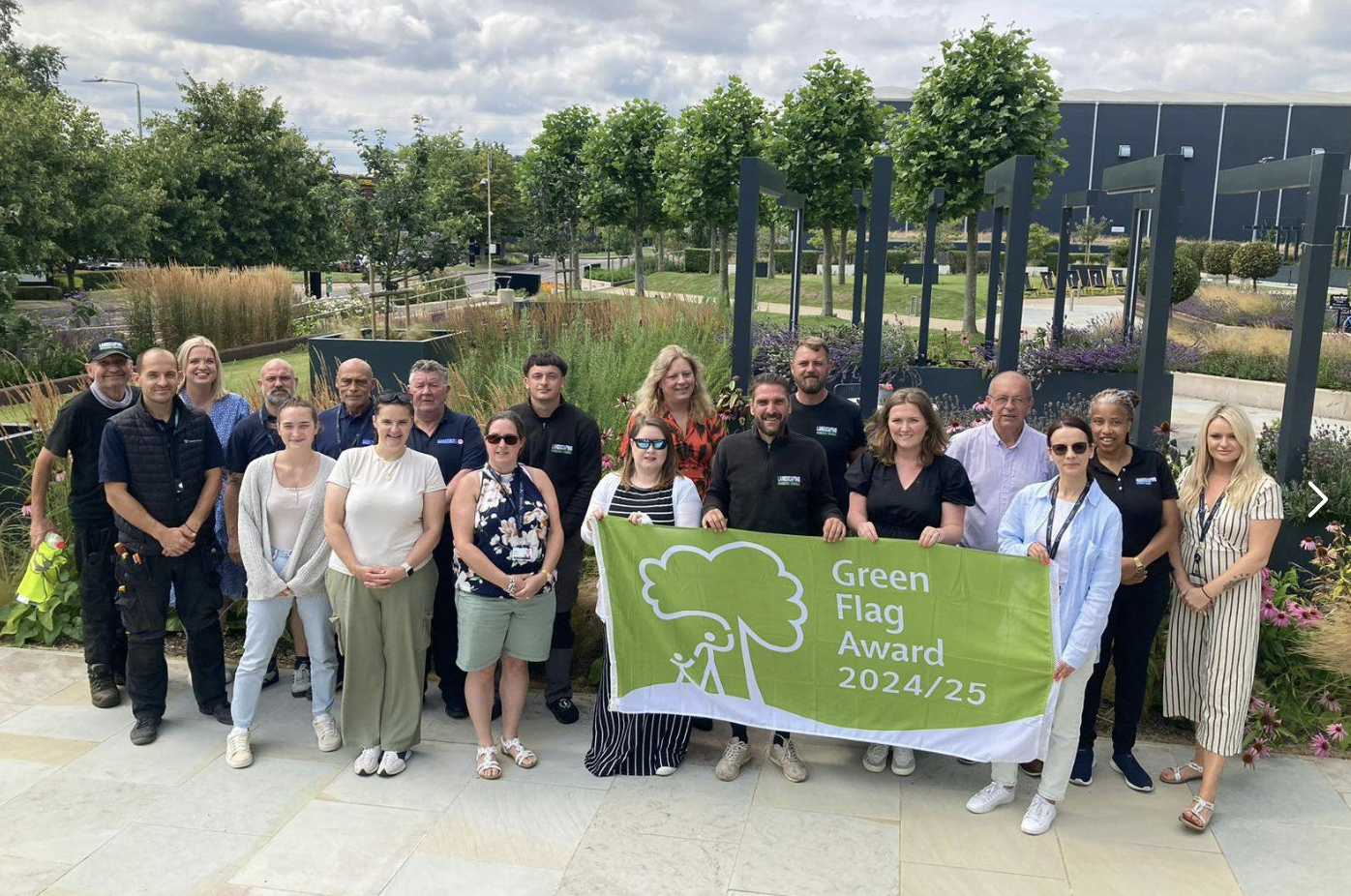 A group of people stand outdoors in a landscaped garden, holding a banner that reads Green Flag Award 2024/25.