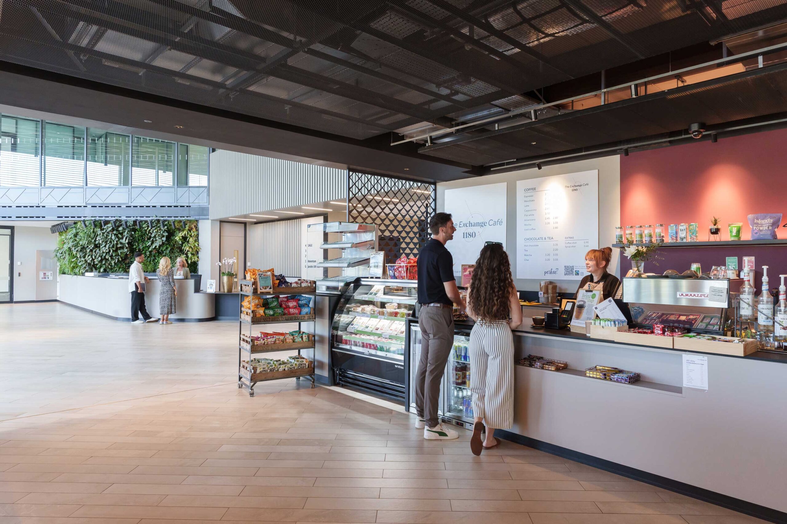 Two people stand at a café counter placing an order whilst a staff member assists them. The café has snacks, a display case with food, and a modern, open interior.