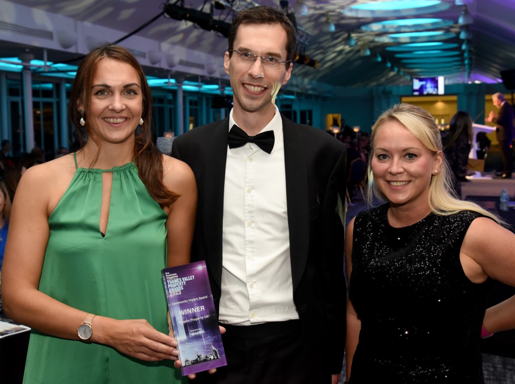 Three people stand together at an indoor event; the person in the centre holds a trophy or award while smiling at the camera.