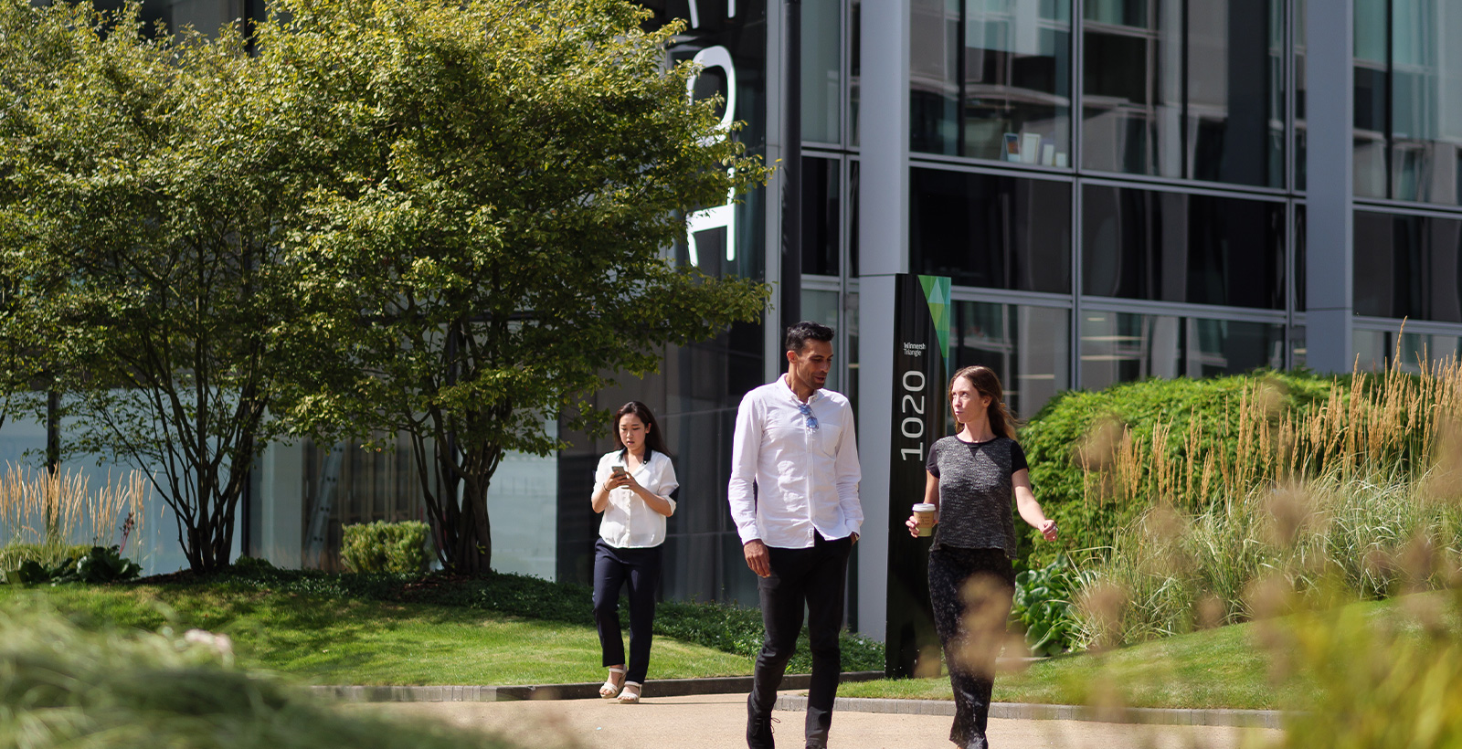 Three people walk outside on a path near a modern office building with glass windows and landscaping.