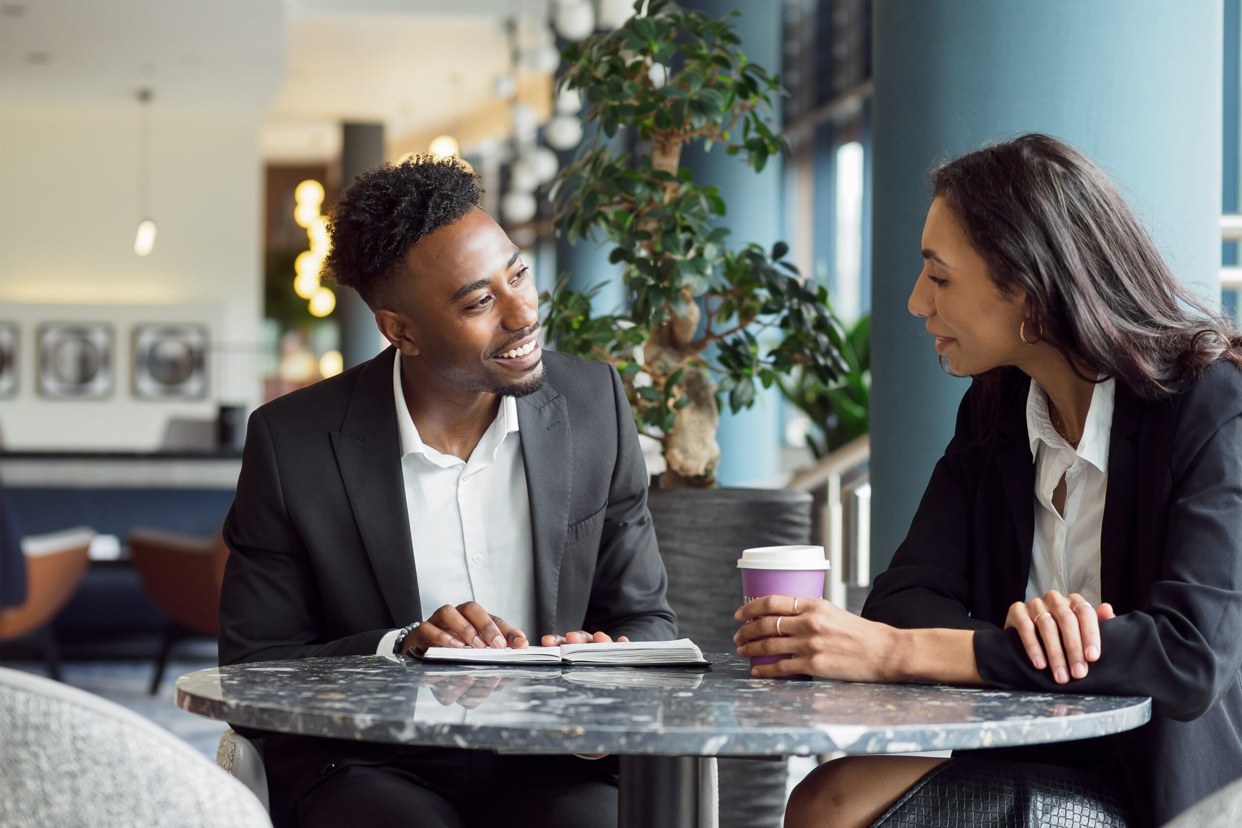 Two people in business attire sit at a round table, talking. One holds a notebook, the other a coffee cup. They appear to be having a professional conversation in a modern office setting.