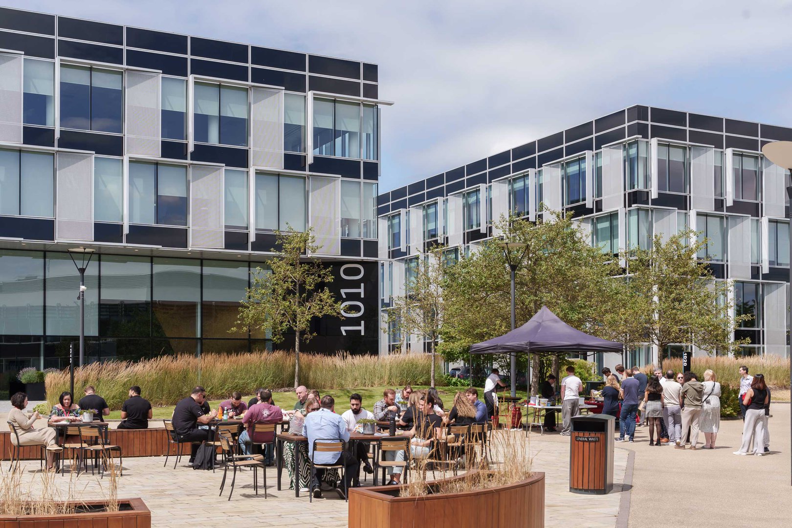 People sit at outdoor tables and stand near a marquee in a modern courtyard with glass office buildings in the background on a sunny day.