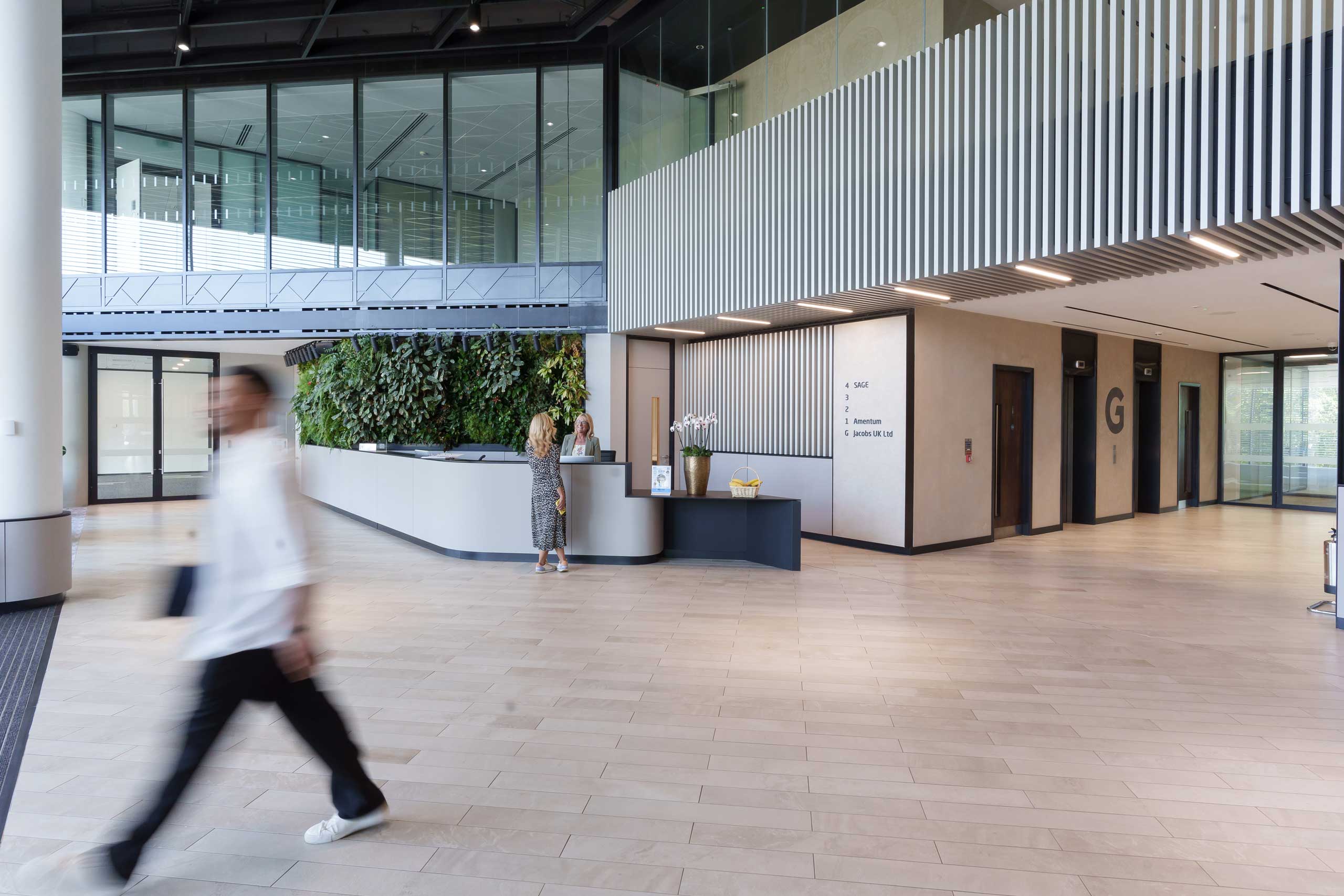 Modern office lobby with a reception desk, green wall, two people talking at the counter, and a person walking by in the foreground.