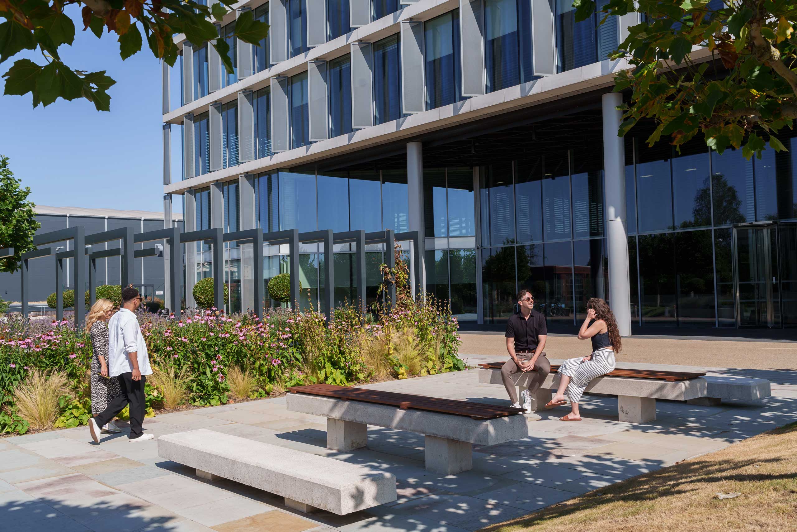 Several people sit and walk outside a modern glass office block with landscaped gardens and benches on a sunny day.