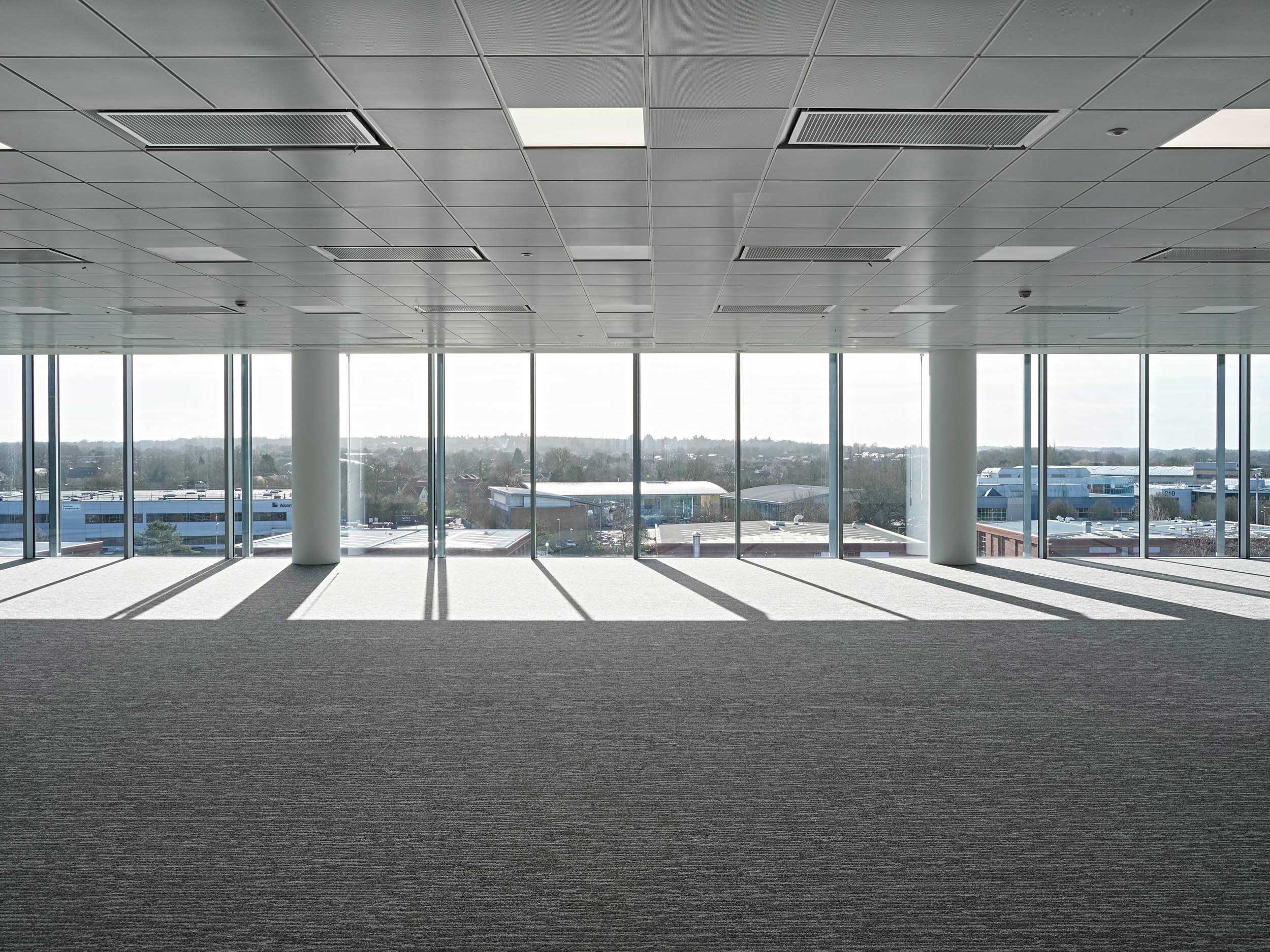 Empty modern office space with large floor-to-ceiling windows, grey carpet, and sunlight casting shadows across the floor. Industrial buildings and trees visible outside.