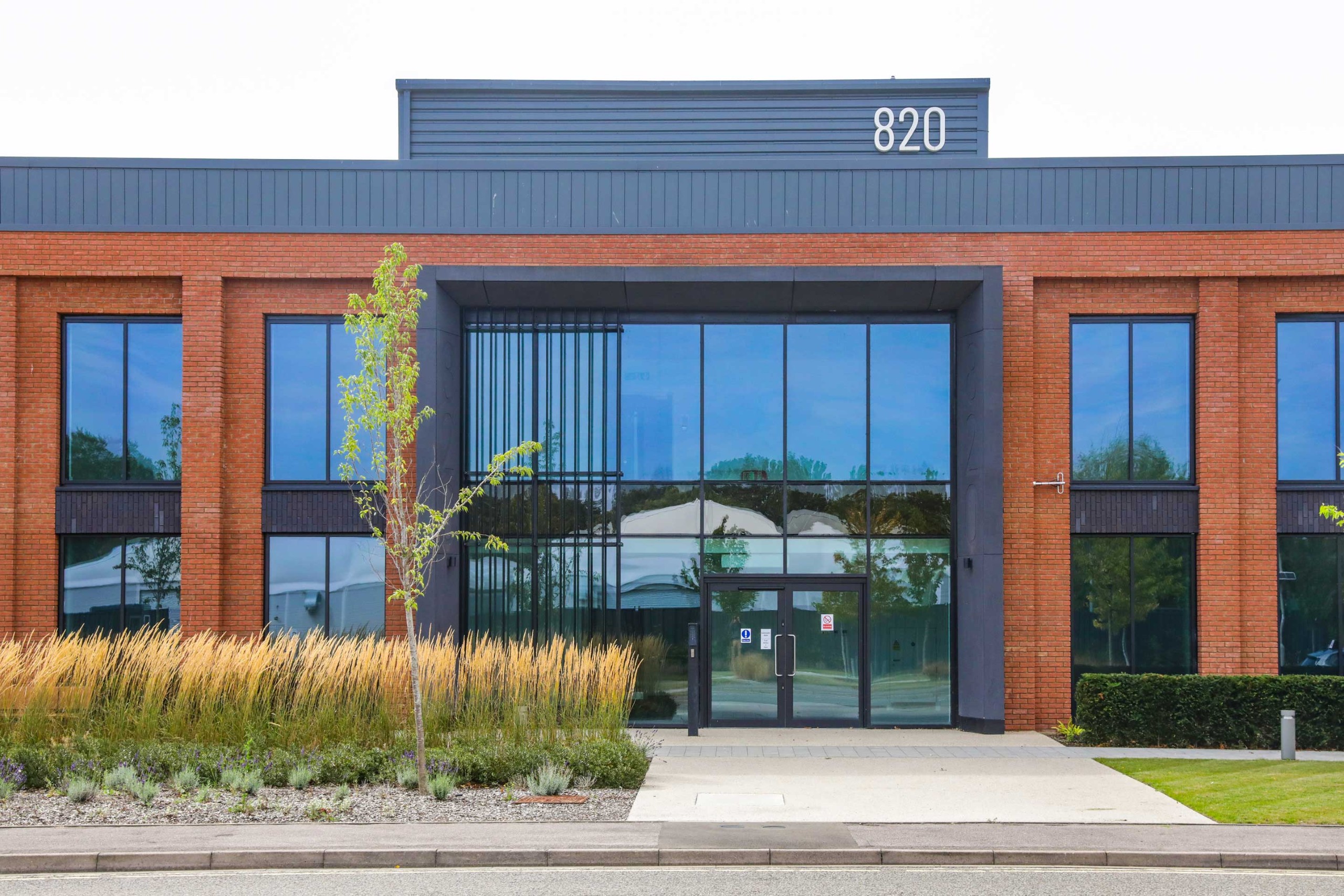 Modern two-storey brick office building with large glass windows, a single tree and landscaped plants in front, and the number 820 displayed at the top.