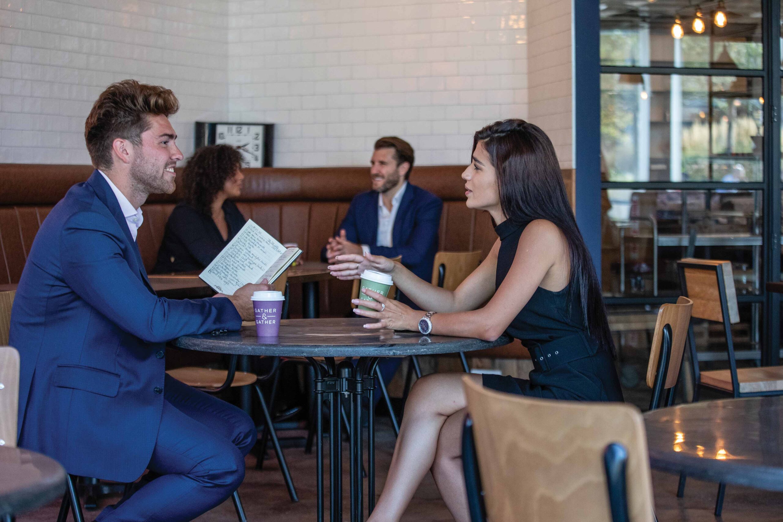 Two people in business attire sit at a café table talking, holding drinks. In the background, another pair also sits and converses.