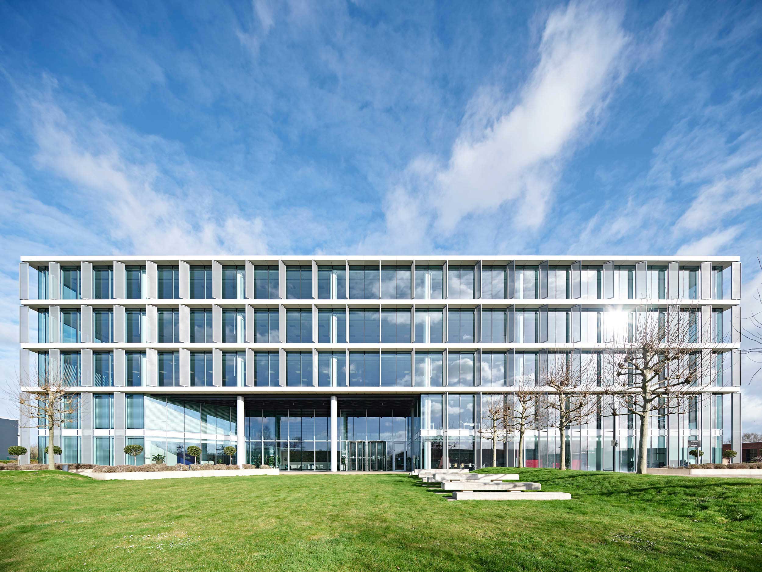 A modern glass office building with a grid-like façade, standing in front of a well-maintained green lawn under a partly cloudy blue sky.