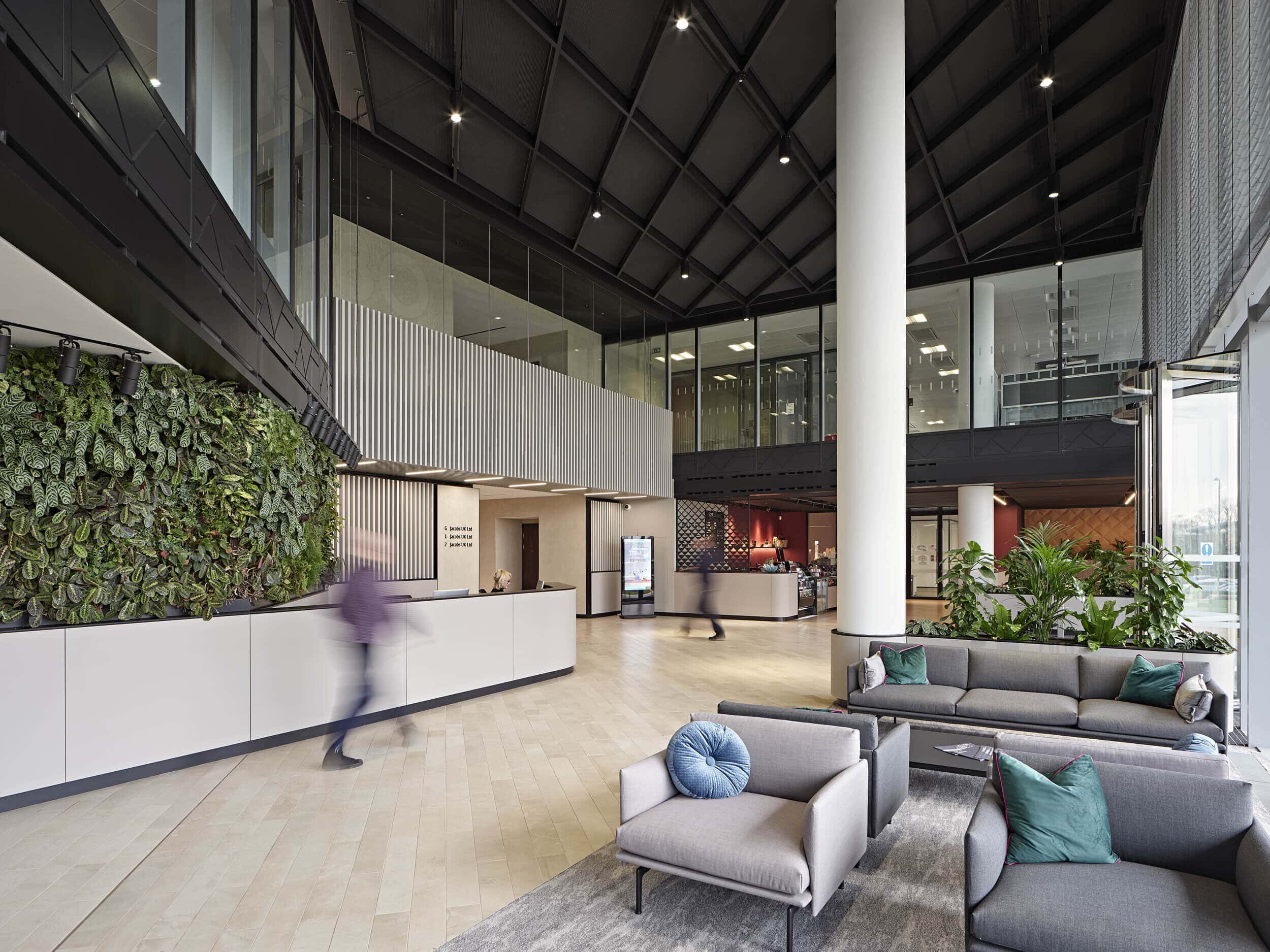 Modern office lobby with high ceilings, a vertical plant wall, reception desk, seating area with grey sofas, large windows, and people walking through the space.