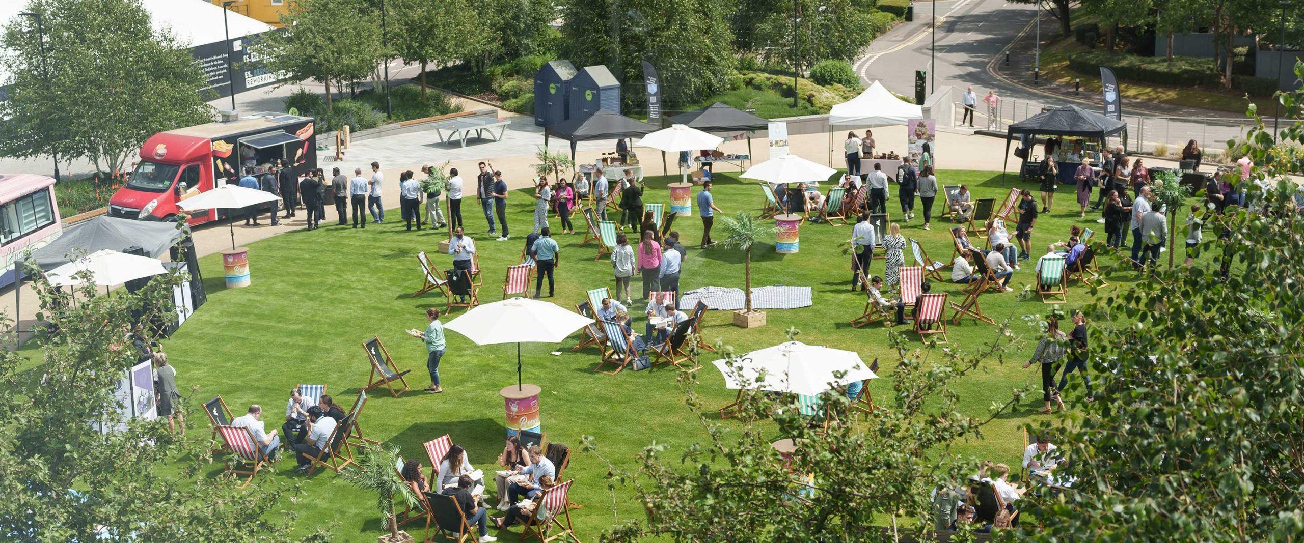 A crowd of people gather on a grassy lawn with deckchairs, umbrellas, and food vans on a sunny day.