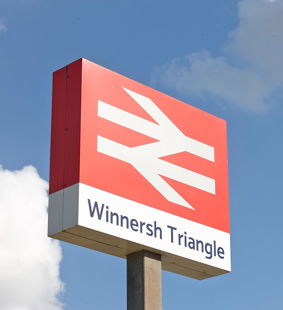 A red and white British Rail logo sign with the text Winnersh Triangle against a blue sky with some clouds.
