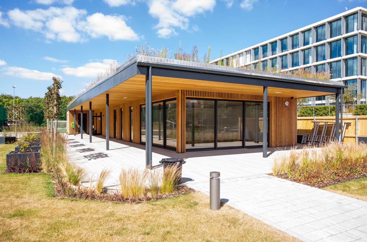 Modern single-storey building with wood panelling, large glass windows, and a green roof, surrounded by landscaping and paved walkway under a blue sky.