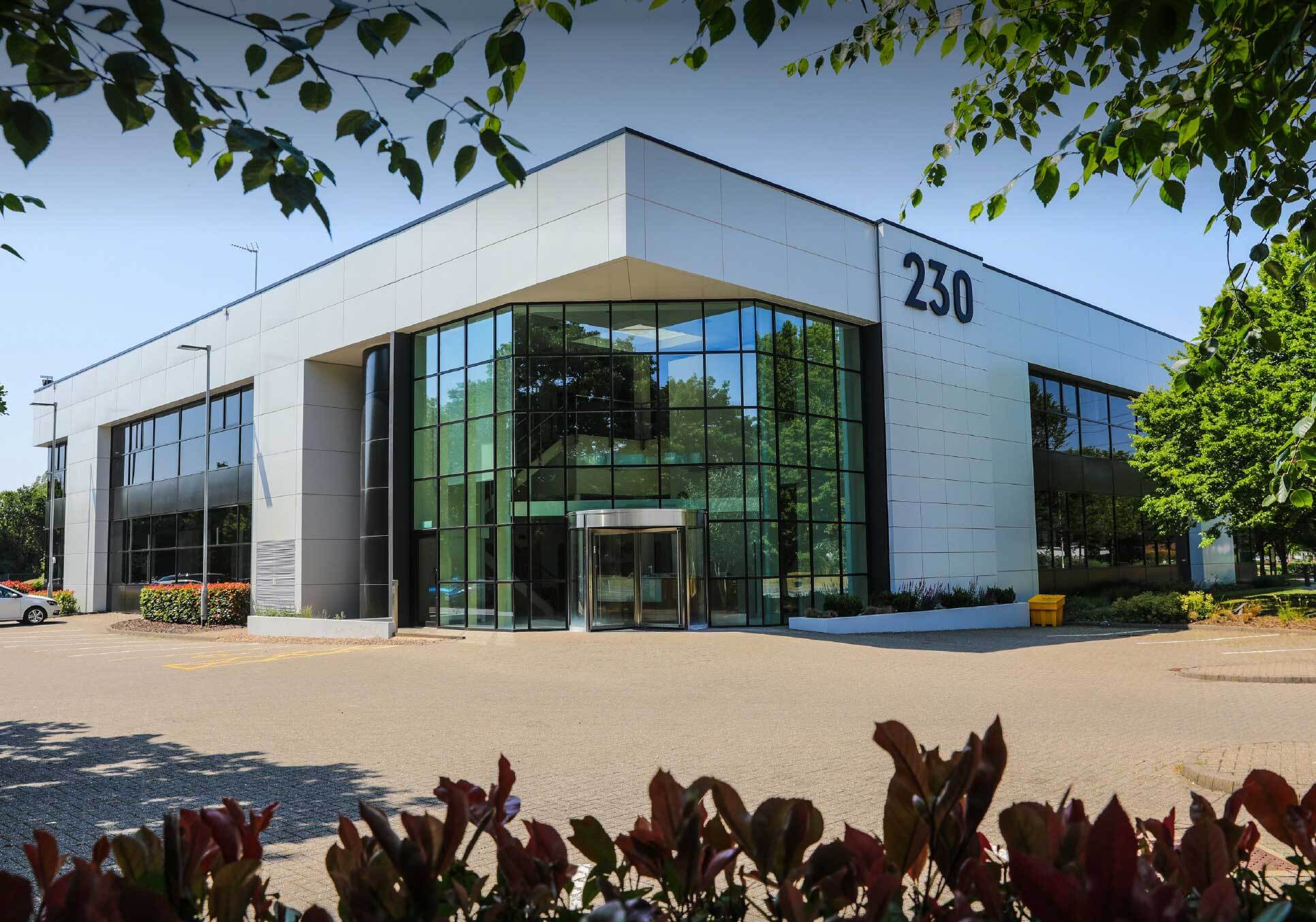 Modern two-storey office building with large glass windows, white exterior panels, and the number 230 displayed on the corner. Trees and bushes frame the entrance area.