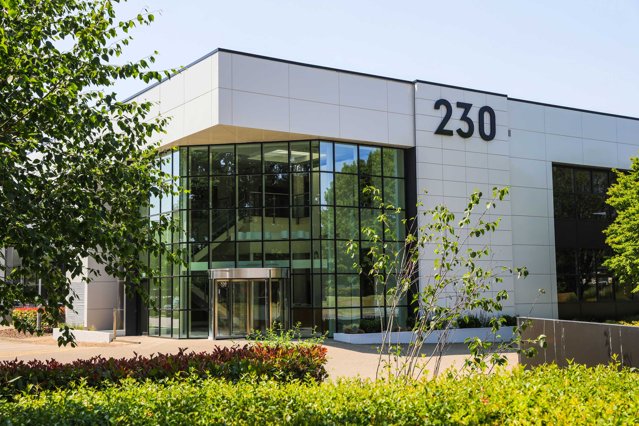 Modern office building with large glass windows and the number 230 displayed on the exterior, surrounded by greenery and landscaping.