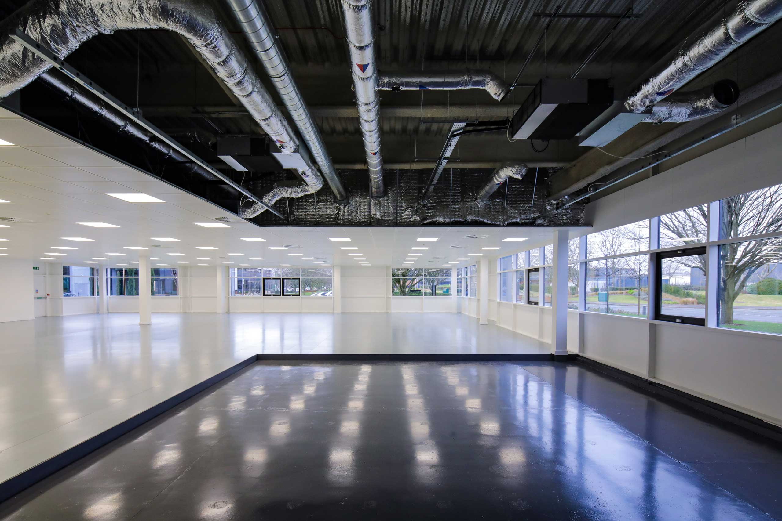 Spacious, empty commercial interior with large windows, exposed ceiling ducts, and a section of glossy dark flooring contrasting with light flooring.