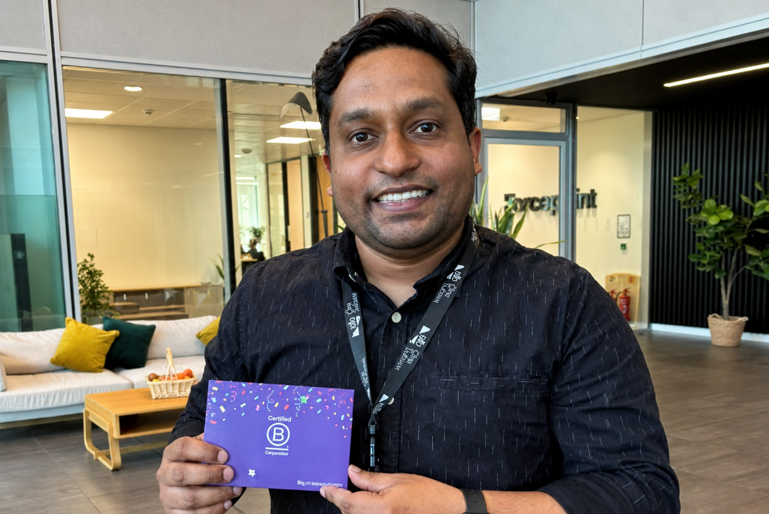 A man stands indoors holding a purple certificate with a Certified B logo, smiling at the camera. Modern office furniture and plants are visible in the background.