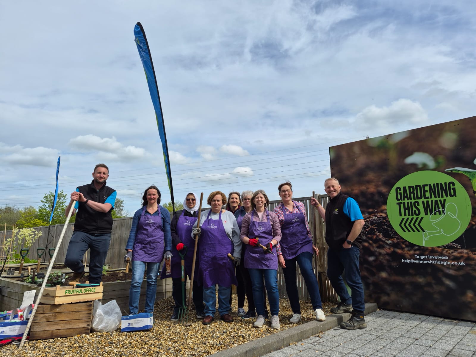 A group of people stand together outdoors by a Gardening This Way sign, some holding gardening tools, with plants and gardening supplies visible around them.